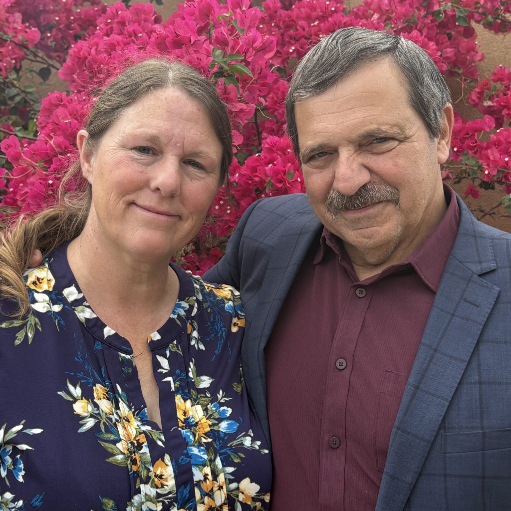 A man and a woman posing together with smiles, set against a backdrop of vibrant pink bougainvillea flowers.